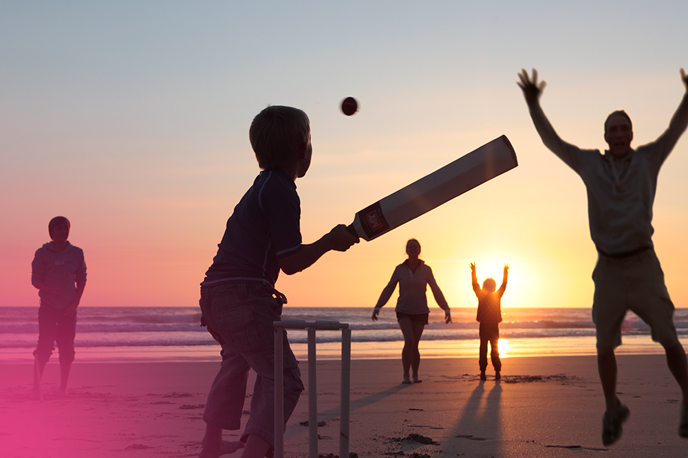 Photo of a young family of five, enjoying a game of cricket on the beach as the sun is setting. The photo has a pink flare in the bottom left corner.