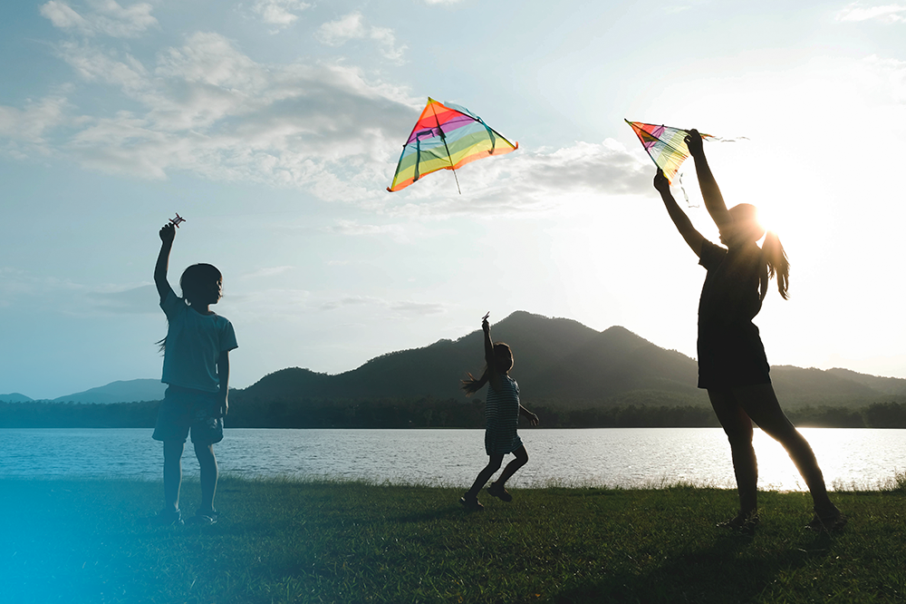 Photo of three young children flying kites in an open field by a lake. The photo has a blue flare in the bottom left corner.