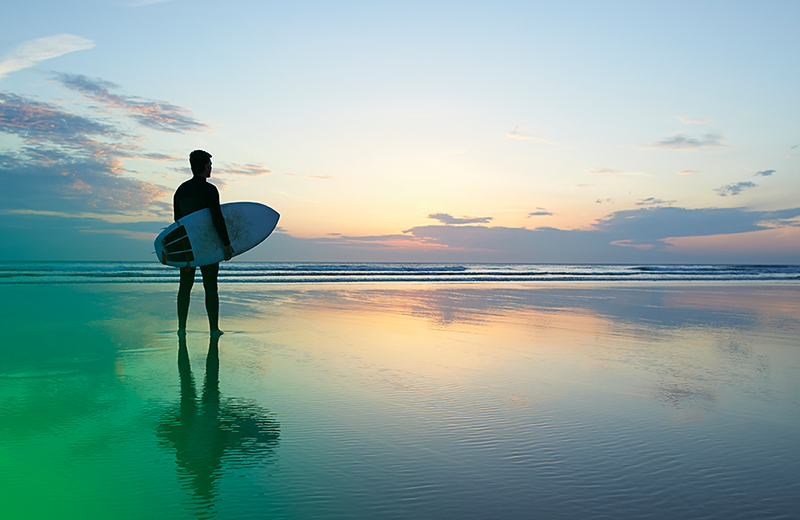 Photo of a man with a surfboard looking out into the ocean and waves. The photo has a green flare in the bottom left corner.