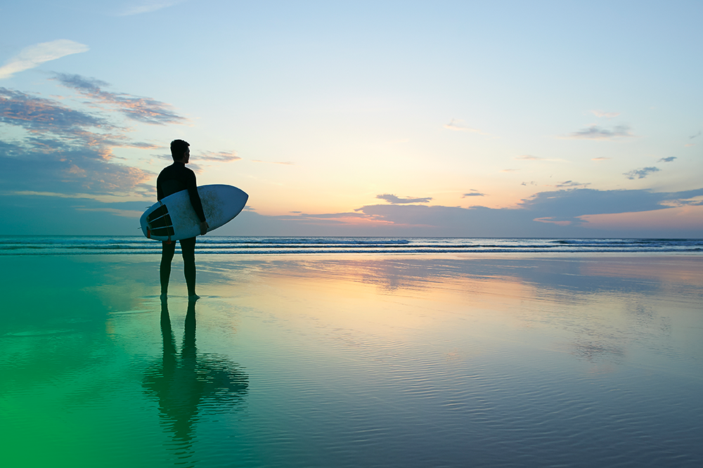 Photo of a man with a surfboard looking out into the ocean and waves. The photo has a green flare in the bottom left corner.