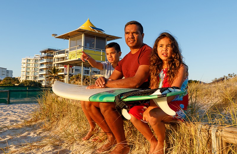 Young family of three sitting on the beach pointing out to the waves, with a surboard resting on their lap.